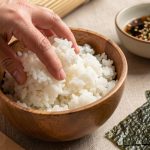 Riz à sushi à la texture équilibrée préparé dans un bol, avec une natte en bambou, une spatule et des feuilles de nori prêtes à rouler.
