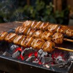 Brasero en acier noir rempli de braises rouges, avec des yakitori de poulet sur doubles brochettes japonaises, éclairés par une lumière chaude du soir.
